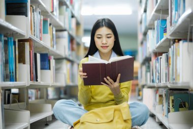 Yerlerde oturmuş kitap okuyan, bir kütüphanede okul projesi için araştırma yapan, günlük giysiler içinde genç bir üniversite öğrencisi. Öğrenim, Eğitim, Kütüphane konsepti.