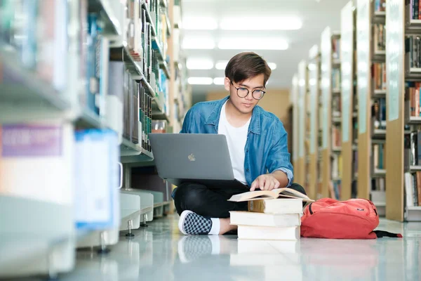 年轻的男大学生 戴着眼镜和休闲装坐在地板上看书 在图书馆用笔记本电脑学习和做研究 电子学习 — 图库照片
