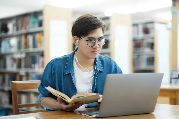 眼鏡やヘッドホンをかけ 机に座ってノートパソコンで読書や読書をしたり 研究や学校のプロジェクトのために図書館でノートパソコンを使用して本を保持する若い男性大学生 — ストック写真