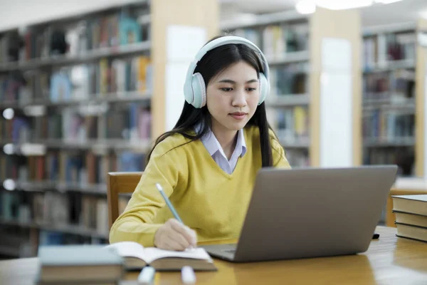 年轻的女大学生 戴着耳机 穿着休闲装坐在写字台前 坐在笔记本上 在图书馆用笔记本电脑学习和做研究 — 图库照片