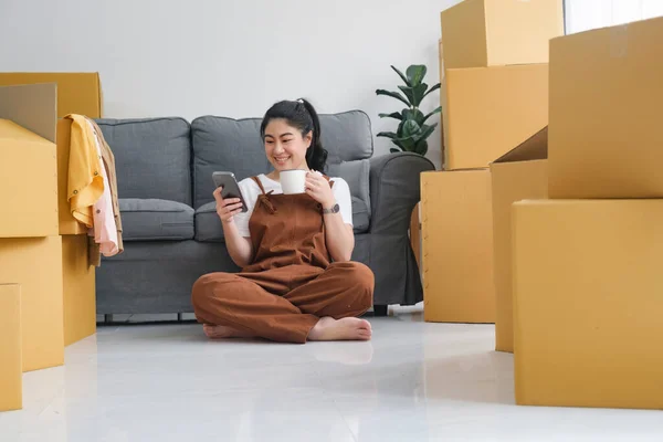 Young woman resting drinking coffee and using mobilephone while moving into new home.