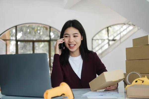 Young asian female online business owner or entrepreneur with smiley face sitting at desk with boxes, laptop, scanner, and taper talking to customer for order using smartphone. Online business, e