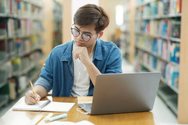 眼鏡をかけ 机に座って勉強したり 本を読んだり 本を読んだり 研究のために図書館でヘッドフォンをしてノートを書き留めたりする若い男性大学生 — ストック写真