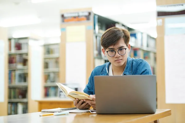 Young male college student wearing eyeglasses and in casual cloths sitting at desk studying, reading book using laptop wearing headphones at library for research or school project. E-Learning concept.