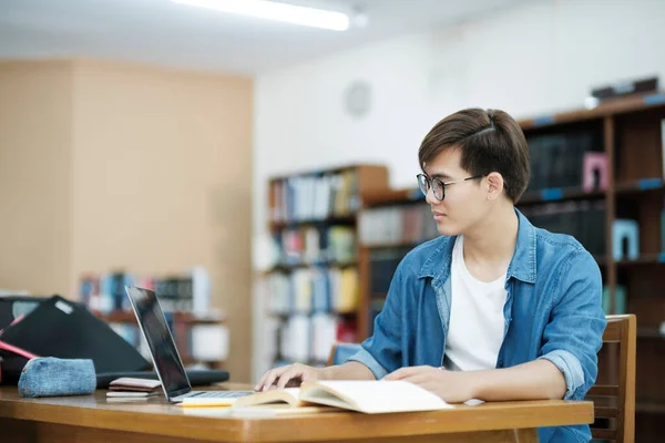 眼鏡をかけた若い男性大学生や 机に座ってノートパソコンを使って本を勉強したり 図書館で本を読んだりして 研究や学校プロジェクトの情報を探したりしています Learning — ストック写真