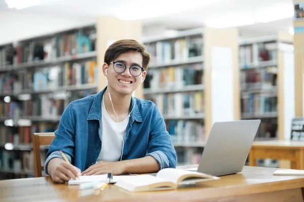 Young male college student wearing eyeglasses and in casual cloths sitting at desk studying, reading book, and writing down notes using laptop wearing headphones looking at camera at library for