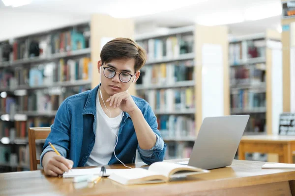 Young male college student wearing eyeglasses and in casual cloths sitting at desk studying, reading book, and writing down notes using laptop wearing headphones at library for research or school
