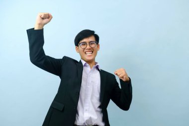 Portriat of asian young smart happy businessman wearing glasses and dressed in suit standing smiling, and flexing at camera to show strength and powerful positive expression with isolated light blue
