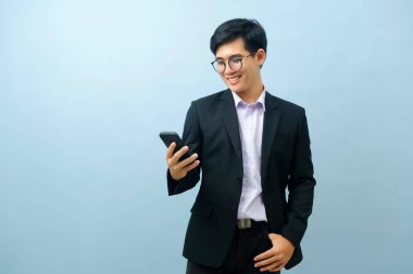 Portrait of young smart asian businessman standing and smiling while using smartphone with light blue isolated background. Business, connection concept.