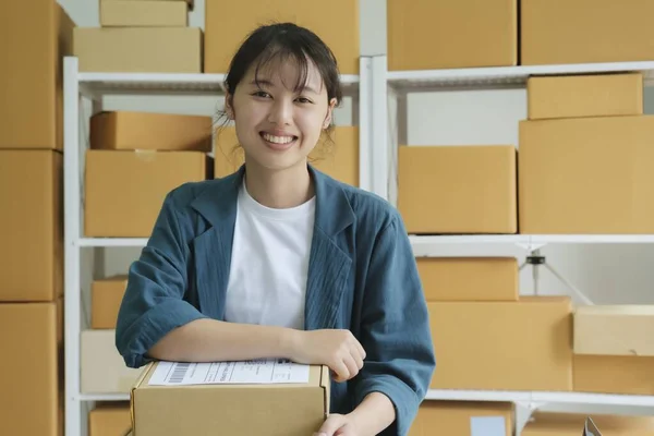 Young female small business entrepreneur, online store owner working ar workplace or warehouse with shelves of cardboard boxes of customers order in the background sitting on desk looking cheerfully