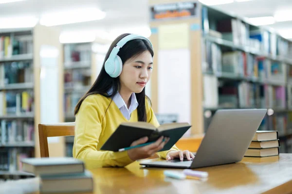 若い女子大生 ヘッドフォンを装着した高校生 机の読書本に座っているカジュアルな服で 図書館で学校プロジェクトのためのラップトップを使用して研究を勉強し 研究を行っています Learning — ストック写真