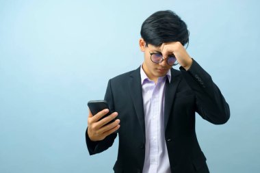 Portrait of young smart asian businessman in eyeglasses standing and talking on mobile phone stressed and frustrated about business with light blue isolated background. Business and connection concept