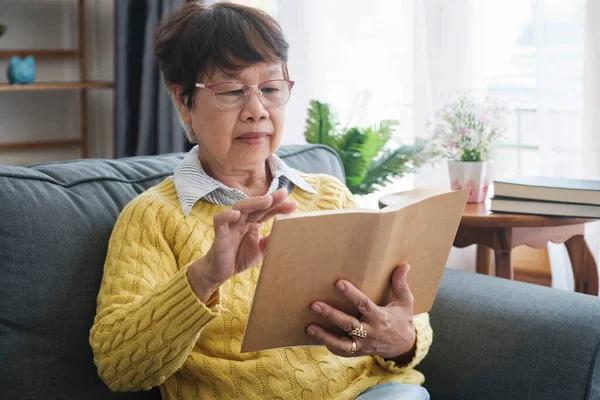 Senior Asian Woman Reading Book Sitting Couch Home Wearing Eyeglasses — Stock Photo, Image
