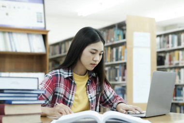 Üniversite Kütüphanesi: Zeki Asyalı üniversite öğrencisi Laptop kullanır, Kağıt için Notlar Yazar, Sınıf ödevi için Çalışma. Odaklanmış öğrenciler öğreniyor, üniversite sınavlarına çalışıyor..