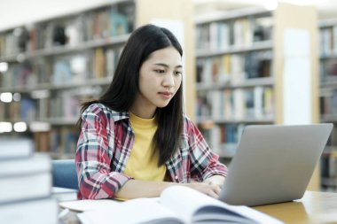 Üniversite Kütüphanesi: Zeki Asyalı üniversite öğrencisi Laptop kullanır, Kağıt için Notlar Yazar, Sınıf ödevi için Çalışma. Odaklanmış öğrenciler öğreniyor, üniversite sınavlarına çalışıyor..