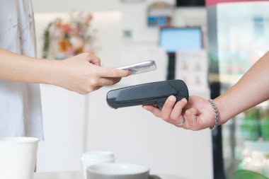 NFC terminali yakınlarında telefonu olan bayan müşteri kahve tezgahında garson bayan baristayla temas kurmadan cep telefonu ödemesi yapıyor, kadın müşteri de kafede pos makinesiyle cep telefonuyla ödeme yapıyor. Tap-to