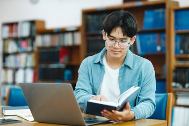 Üniversite Kütüphanesi: Zeki Asyalı üniversite öğrencisi Laptop kullanır, Kağıt için Notlar Yazar, Sınıf ödevi için Çalışma. Odaklanmış öğrenciler öğreniyor, üniversite sınavlarına çalışıyor..