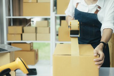 Çevrimiçi işletme sahibi müşteriye teslimat için ürün hazırlar. Çevrimiçi girişimci paket kutusunu mühürlemek için koli bandı kullanıyor. Küçük işletme sahibi, KOBİ kavramı.