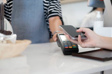 NFC terminali yakınlarında telefonu olan bayan müşteri kahve tezgahında garson bayan baristayla temas kurmadan cep telefonu ödemesi yapıyor, kadın müşteri de kafede pos makinesiyle cep telefonuyla ödeme yapıyor. Tap-to