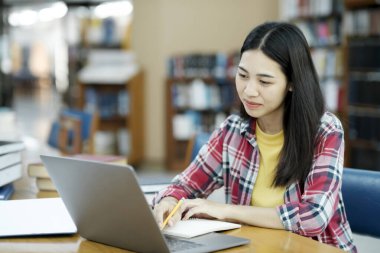 Üniversite Kütüphanesi: Zeki Asyalı üniversite öğrencisi Laptop kullanır, Kağıt için Notlar Yazar, Sınıf ödevi için Çalışma. Odaklanmış öğrenciler öğreniyor, üniversite sınavlarına çalışıyor..