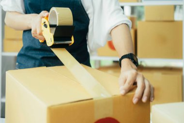 Çevrimiçi işletme sahibi müşteriye teslimat için ürün hazırlar. Çevrimiçi girişimci paket kutusunu mühürlemek için koli bandı kullanıyor. Küçük işletme sahibi, KOBİ kavramı.