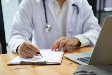 Tıbbi kayıtları dolduran pratisyen hekimin elleri. Beyaz önlüklü doktor iş yerinde dizüstü bilgisayarla evrak işleriyle uğraşıyor, notlar yazıyor, evrak hazırlıyor, raporlar hazırlıyor, reçete yazıyor..
