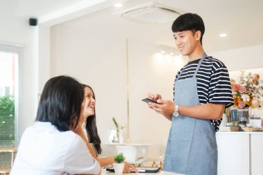 Önlüklü neşeli bir garson, sipariş verirken ve müşterilerle konuşurken kahve dükkanı ziyaretçileri, arkadaş canlısı profesyonel servis elemanı önlüğü giyme menü seçimi, iyi müşterilere servis yapma.