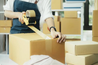 Çevrimiçi işletme sahibi müşteriye teslimat için ürün hazırlar. Çevrimiçi girişimci paket kutusunu mühürlemek için koli bandı kullanıyor. Küçük işletme sahibi, KOBİ kavramı.