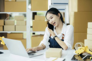 Küçük işletme sahipleri, başlangıç ve e-ticaret konsepti. Gülümseyen Asyalı kadın müşteriyle telefonda konuşuyor, sevkiyat siparişlerini paketlerken telefonlara cevap veriyor, internetten alışveriş yapıyor, dizüstü bilgisayar kullanıyor.