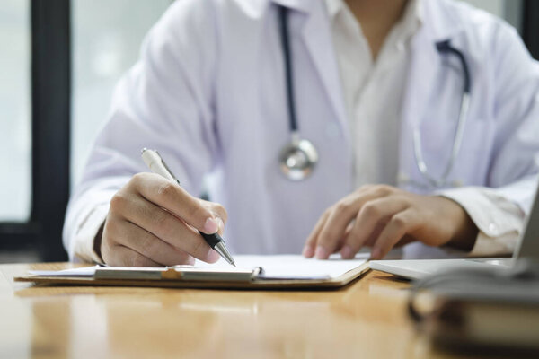 Hands of general practitioner filling paper medical records. Doctor in white coat doing paperwork at workplace with laptop, writing notes, preparing documents, reports, prescription.