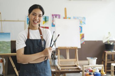 Genç sanatçı kadın, atölyesinde ya da sanat galerisinde kameraya gülümseyen fırça ve fırçaları tutan bir önlük giyiyor. Hobi ve yaşam tarzı
