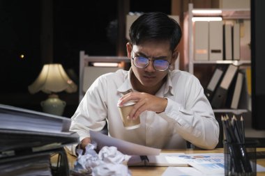 Genç Asyalı işadamı yorgun bir ofis çalışanı olarak masa başında bilgisayar kullanıyor ve fazla mesai yapıyor..