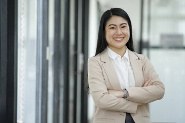 Profesyonel ve kendine güvenen bir iş kadını kameraya gülümsüyor. Masasının başında dizüstü bilgisayarla modern bir ofiste duruyor..