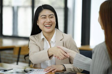 İki gülümseyen iş kadını ofiste masanın üzerinden el sıkışıyor. Başarılı bir anlaşmayı ya da ortaklığı simgeliyor..