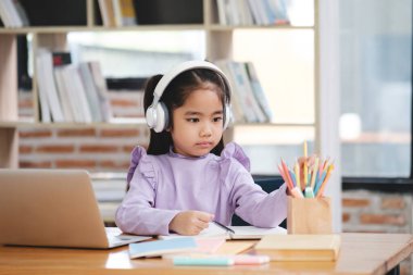 Kulaklık takan genç bir kız bir dizüstü bilgisayar ve bir kutu renkli kalemle bir masada oturuyor. İşine odaklanmış durumda, muhtemelen ders çalışıyor ya da ev ödevi yapıyor. Sahne sessiz bir yer öneriyor.