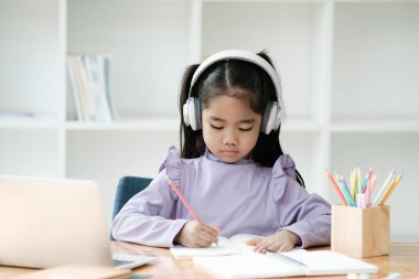 Genç bir kız dizüstü bilgisayarı ve not defteriyle bir masada oturuyor. Kulaklık takıyor ve defterine yazıyor.