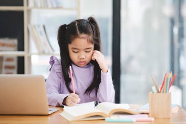 Genç bir kız bir dizüstü bilgisayar ve bir kitapla masada oturuyor. O bir kalemle yazıyor ve işine odaklanmış durumda.