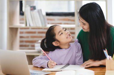 Bir kadın genç bir kıza ev ödevinde yardım ediyor. Kız mor bir gömlek giyiyor ve bir dizüstü bilgisayar ve bir not defteriyle bir masada oturuyor. Kadın kızların elini tutuyor.