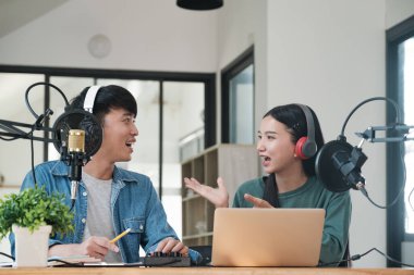 İki kişi mikrofonda konuşuyor. İçlerinden biri kulaklık takıyor. Diğer kişi yeşil gömlek giyiyor.