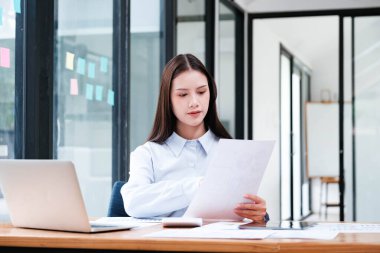 Ofis masasındaki bir belgeyi dikkatle inceleyen odaklanmış bir kadın profesyonel. Yanında bir laptop ve akıllı telefon var..