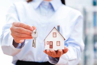Emlak Temsilcisi Ev Anahtarları ve Model Ev 'i sunar, Yeni Ev Sahipliği ve Gayrimenkul İşlemlerinin Keyfi Vurgulanır