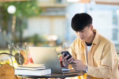 Bir üniversite öğrencisi laptopunda çalışırken düşünceye dalıp kampüsün sakinliğiyle çevrili bir açık hava masasında oturuyor..