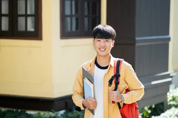 一个快乐而满足的年轻人 拿着笔记本电脑和书本 站在大学校园外面 体现了大学生活的乐观情绪 图库照片