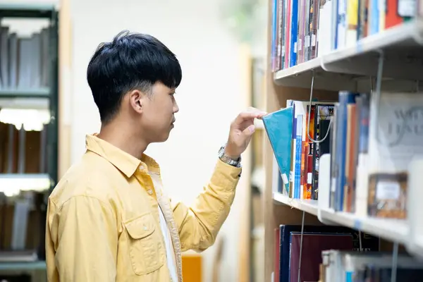 一个穿着黄色衬衫的男人正在看图书馆架子上的一本书 他在摸一本蓝色封面的书 图库图片