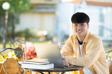 Bir üniversite öğrencisi laptopunda çalışırken düşünceye dalıp kampüsün sakinliğiyle çevrili bir açık hava masasında oturuyor..