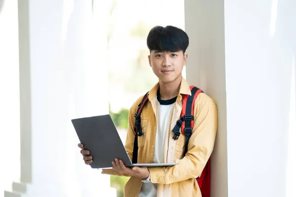 一个面带笑容的男生 自信地拿着笔记本电脑 站在学校的走廊里 准备上课 图库图片