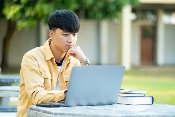 대학생은 자신의 노트북에 공부하는 생각에 캠퍼스의 평온에 둘러싸인 테이블에 스톡 이미지
