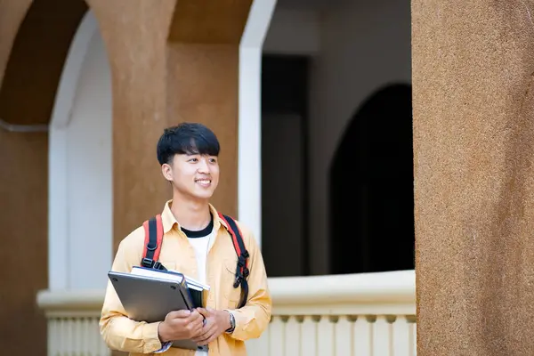 一个心满意足的大学生拿着笔记本电脑和书本在校园里走来走去 流露出一种学习的准备感和热情 免版税图库图片