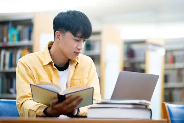 一个年轻人坐在图书馆的桌子旁 一边看书 一边用笔记本电脑 专注和集中的概念 因为男人从事他的研究 图书馆的环境让人觉得很安静 图库图片