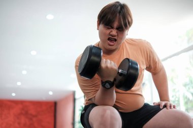 Kilolu bir adam bankta oturuyor, spor salonunda ağır bir halteri kaldırırken yüzünü buruşturuyor..